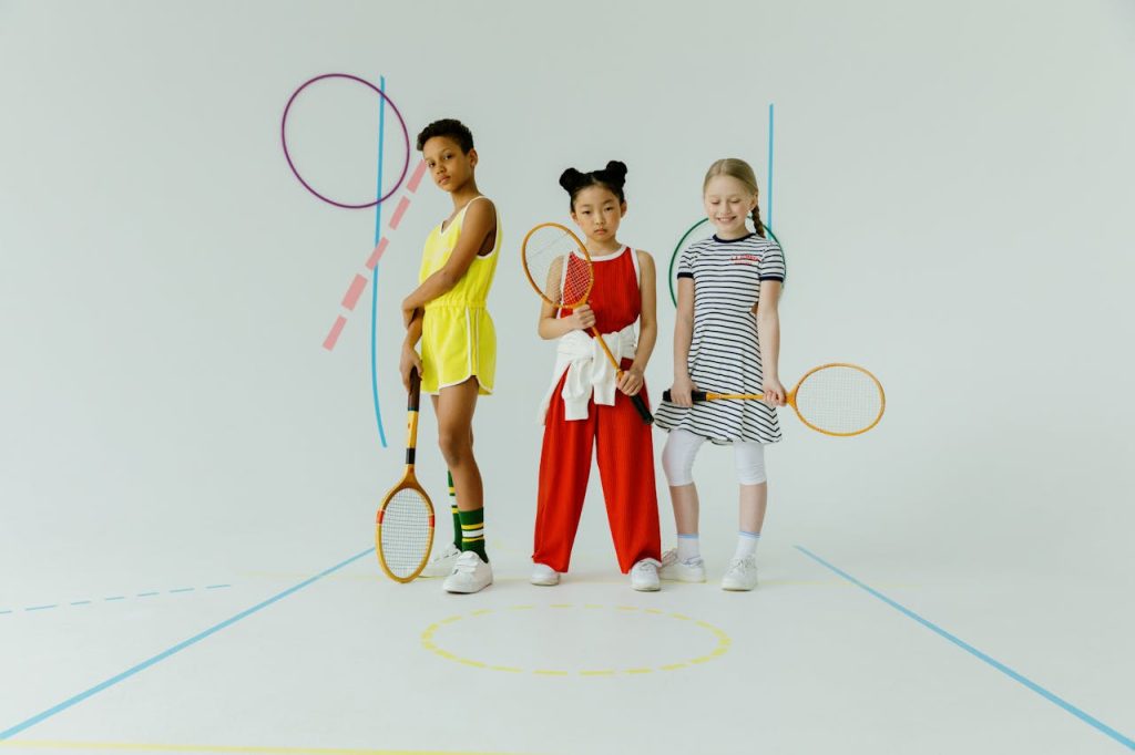 Fun photo of three diverse children in colorful sportswear holding badminton rackets in a studio setting.